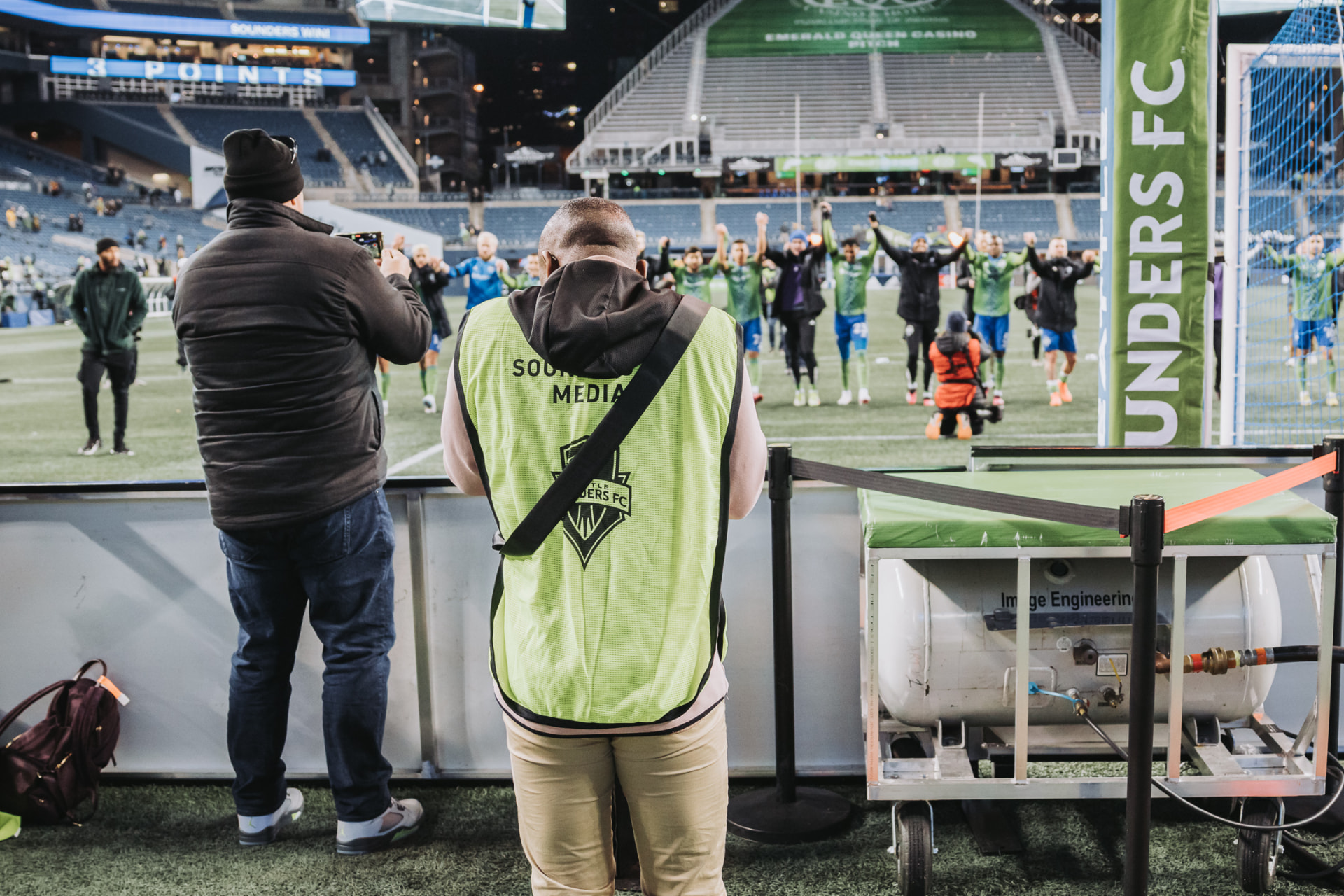Nathan Berg works pitchside in a Sounders FC media vest while players acknowledge supporters after the match.