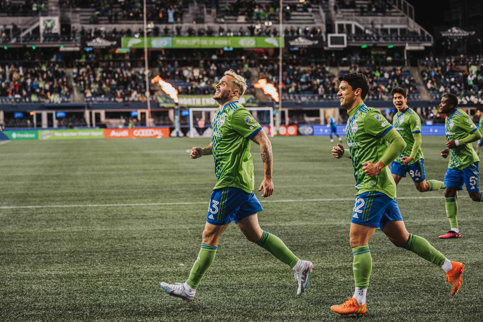 Seattle Sounders players celebrate a goal in green kit as flames rise pitchside in the stadium.