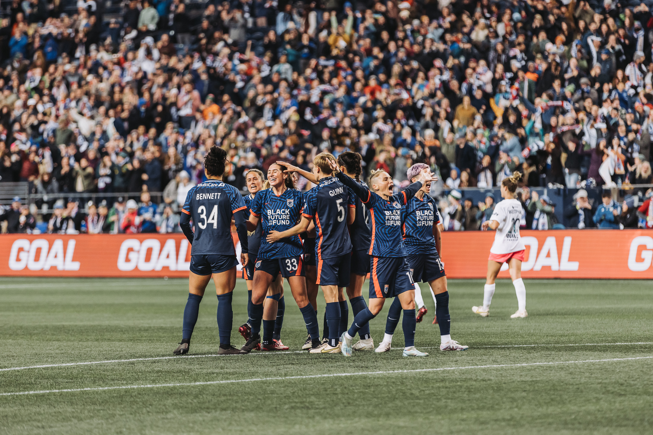 OL Reign players gather to celebrate a goal in front of a packed stand, with the pitchside GOAL board lit behind them.