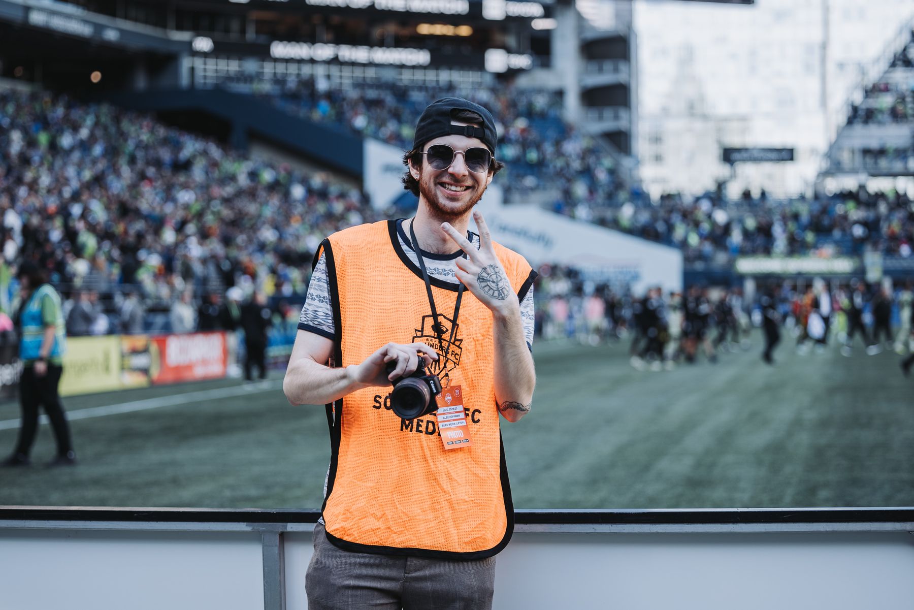 Alec Hoffman stands pitchside holding his camera, his match photo credential hanging from a lanyard.