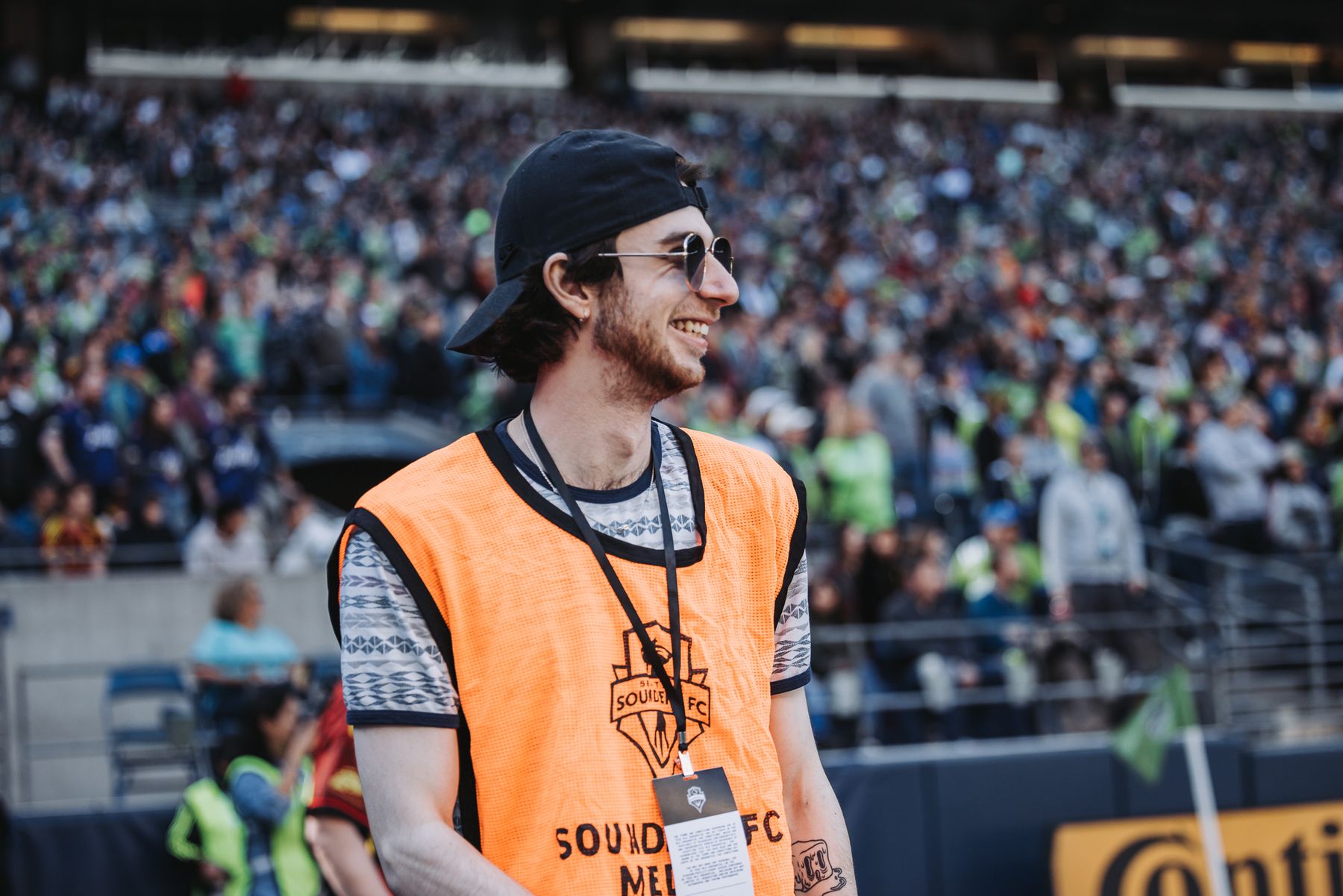Alec Hoffman smiles and looks off to the side in a Sounders FC media vest, with the crowd out of focus behind him.