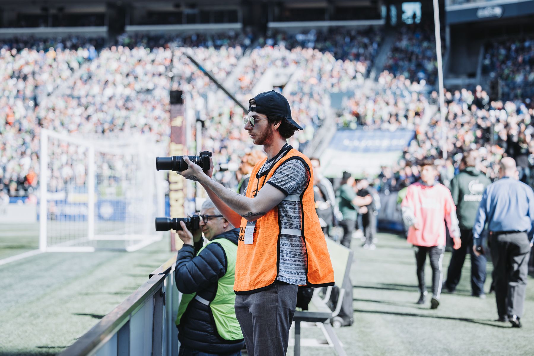 Alec Hoffman stands at the edge of the pitch in an orange media vest, reviewing his camera as the stadium fills behind him.