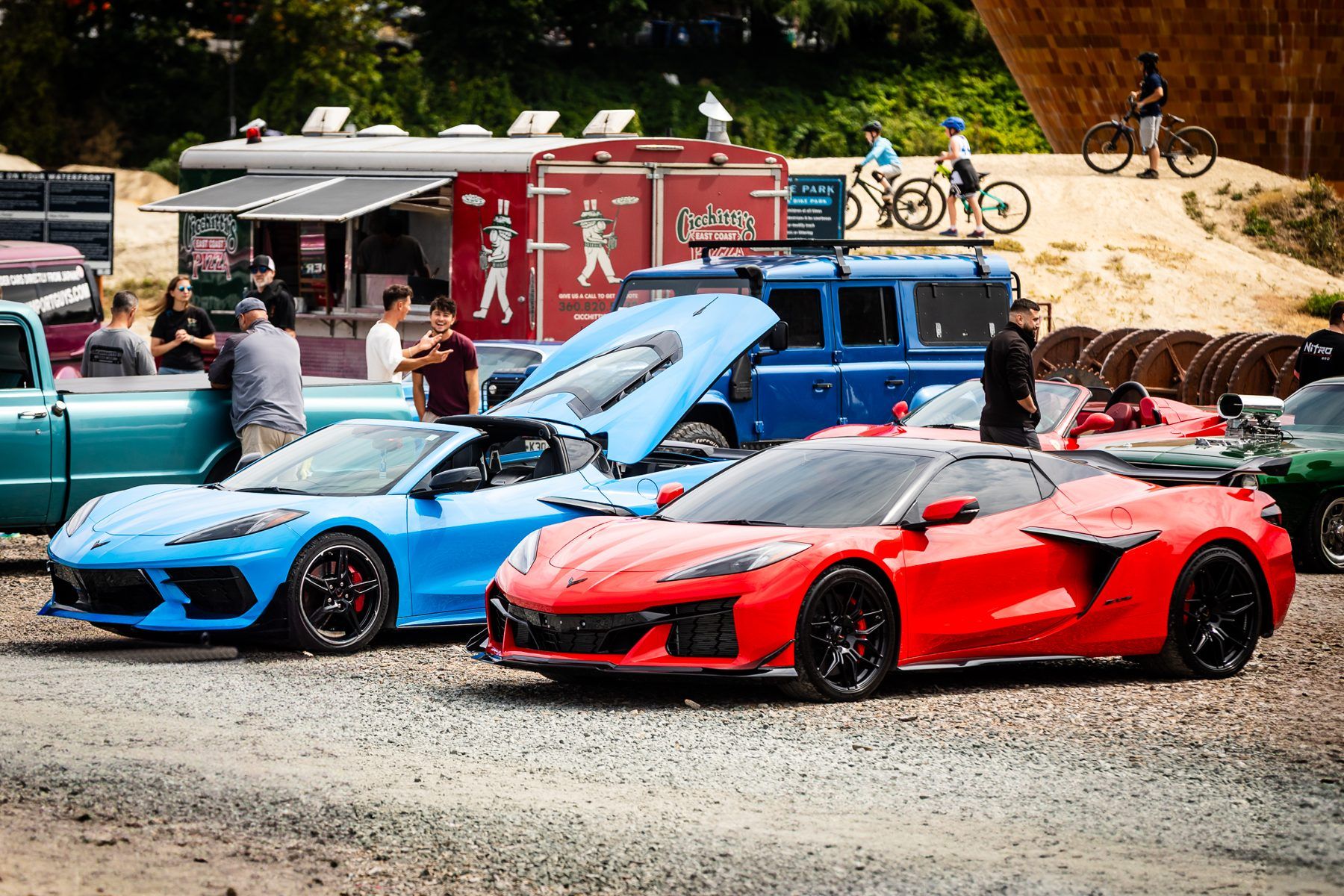 A blue and a red sports car parked side by side on gravel near a food truck at a car show.