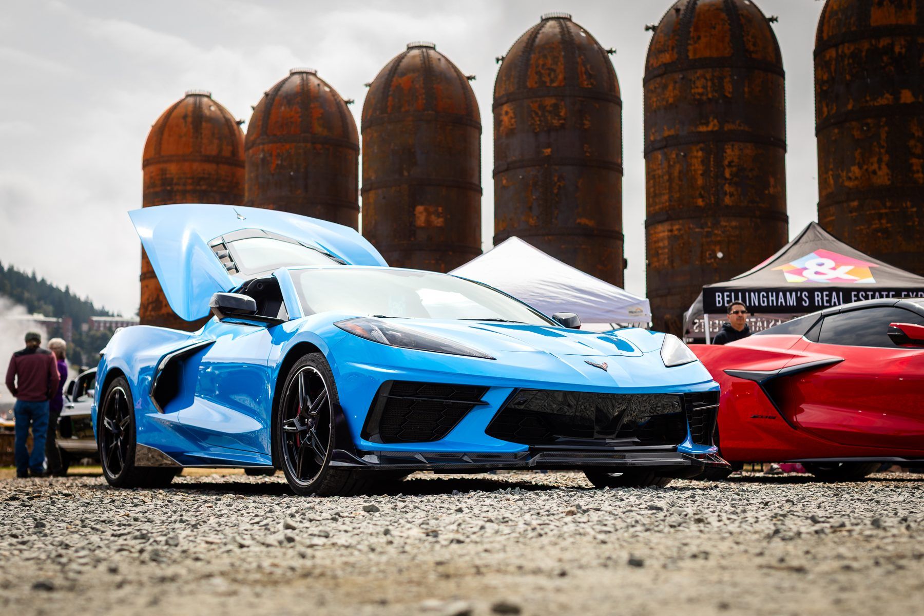 A blue sports car with its hood raised in front of tall rusted waterfront tanks.