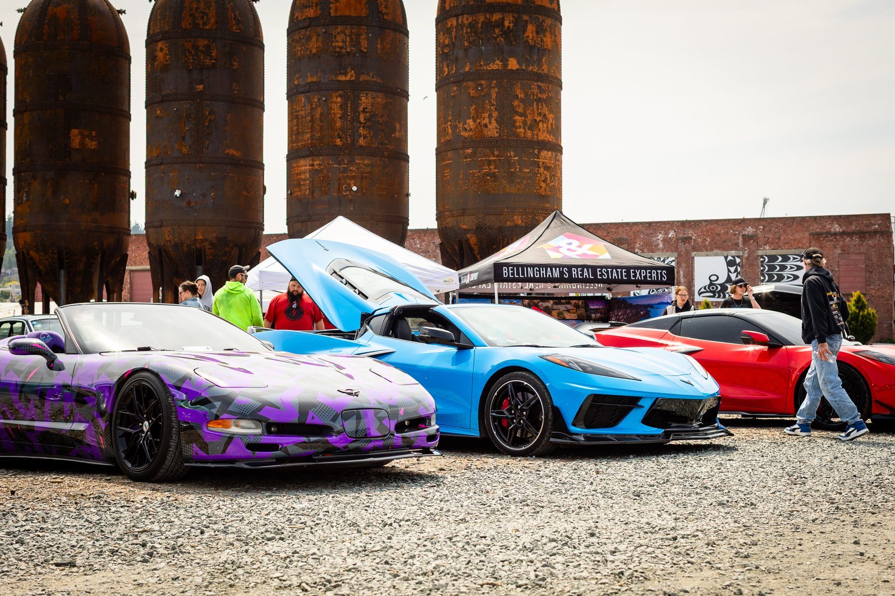 A purple wrapped car, a blue car and a red car lined up on gravel under rusted tanks.