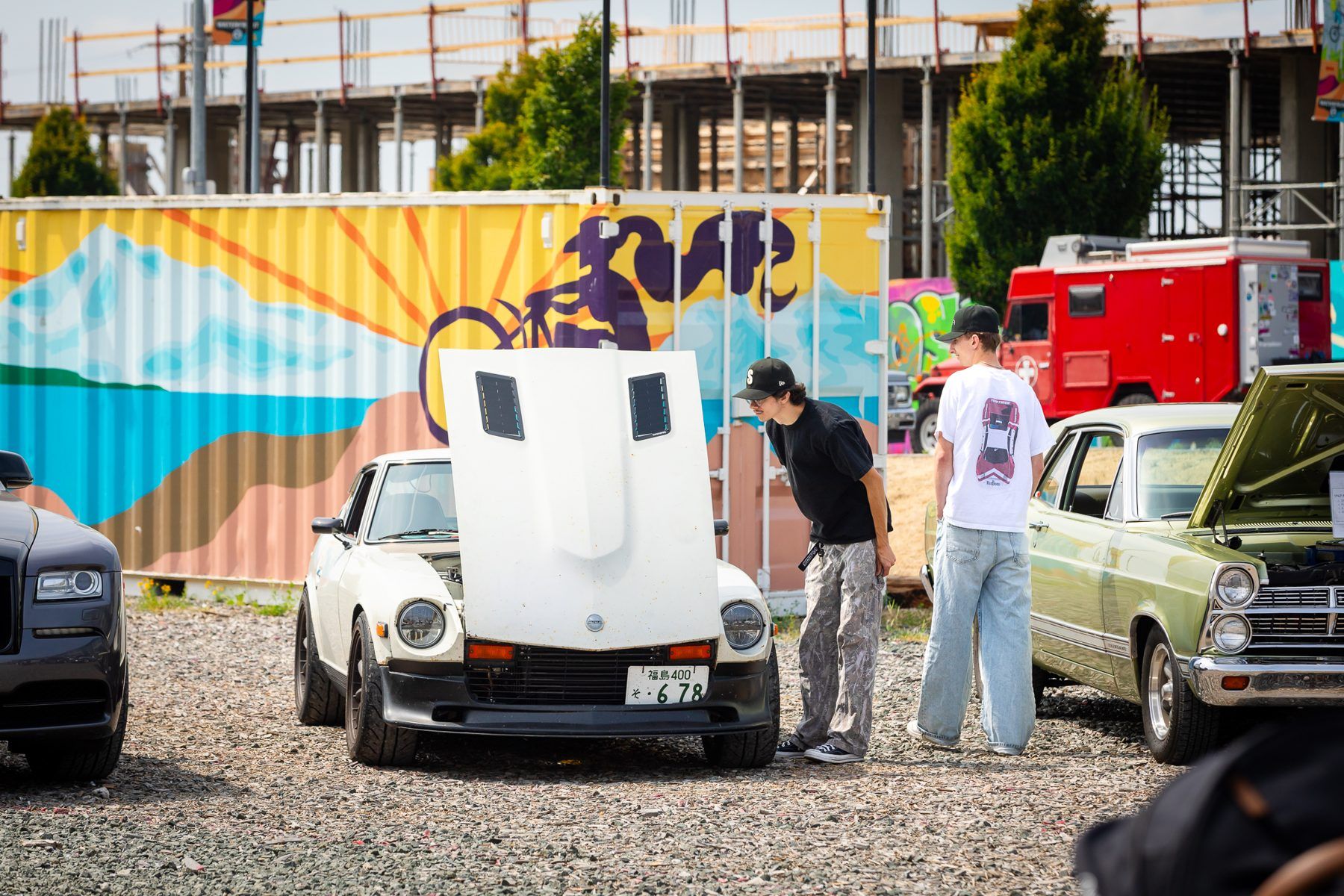 Two people look at a white classic sports car with its hood up beside a painted shipping container.