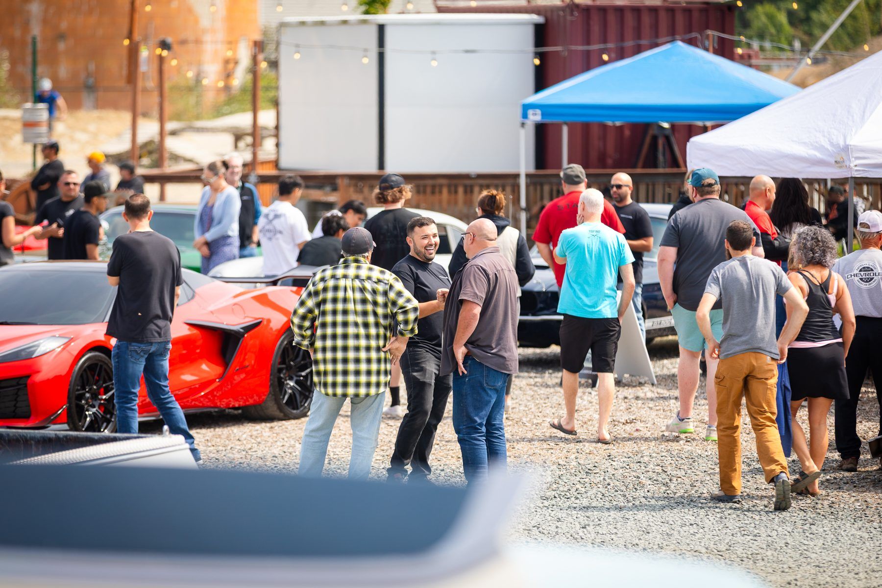 A crowd of visitors talking and walking between parked cars on a gravel lot at a car show.