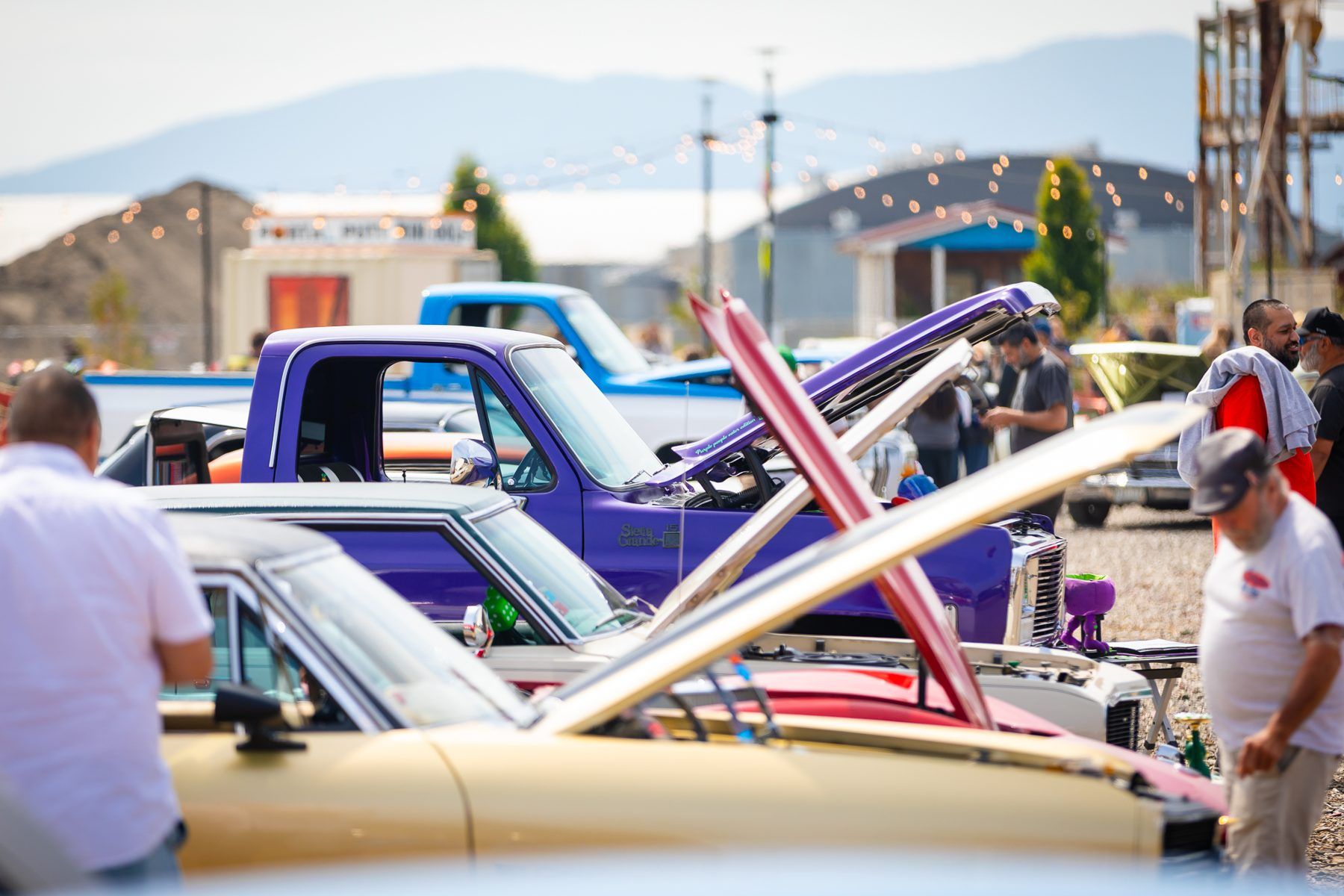 A row of classic trucks and cars with open hoods, with mountains behind the show field.