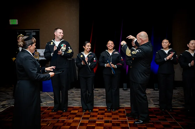 Navy sailors in dress uniforms clap on stage at a formal ball