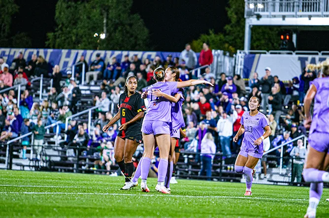 Women's soccer players in purple celebrate a goal in front of a full stand at night