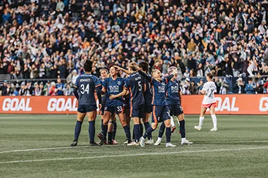 Soccer players in navy celebrate in front of a packed stadium crowd
