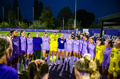 Women's soccer players in purple huddle on the field at dusk