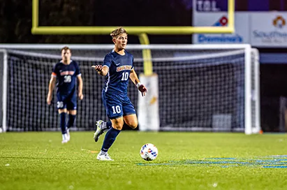A soccer player in navy dribbles toward goal under stadium lights