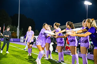Women's soccer players in purple high five teammates on the field at night