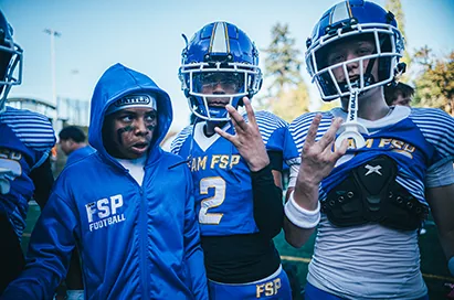 Football players in blue throw up hand signs on the sideline