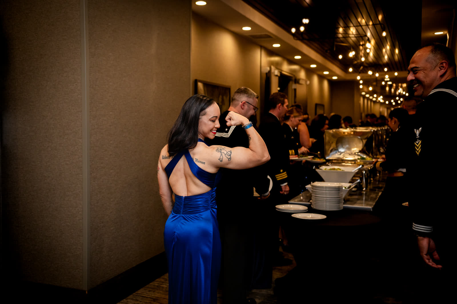 A guest in a blue gown flexes and laughs beside the buffet line during the Navy Ball reception.