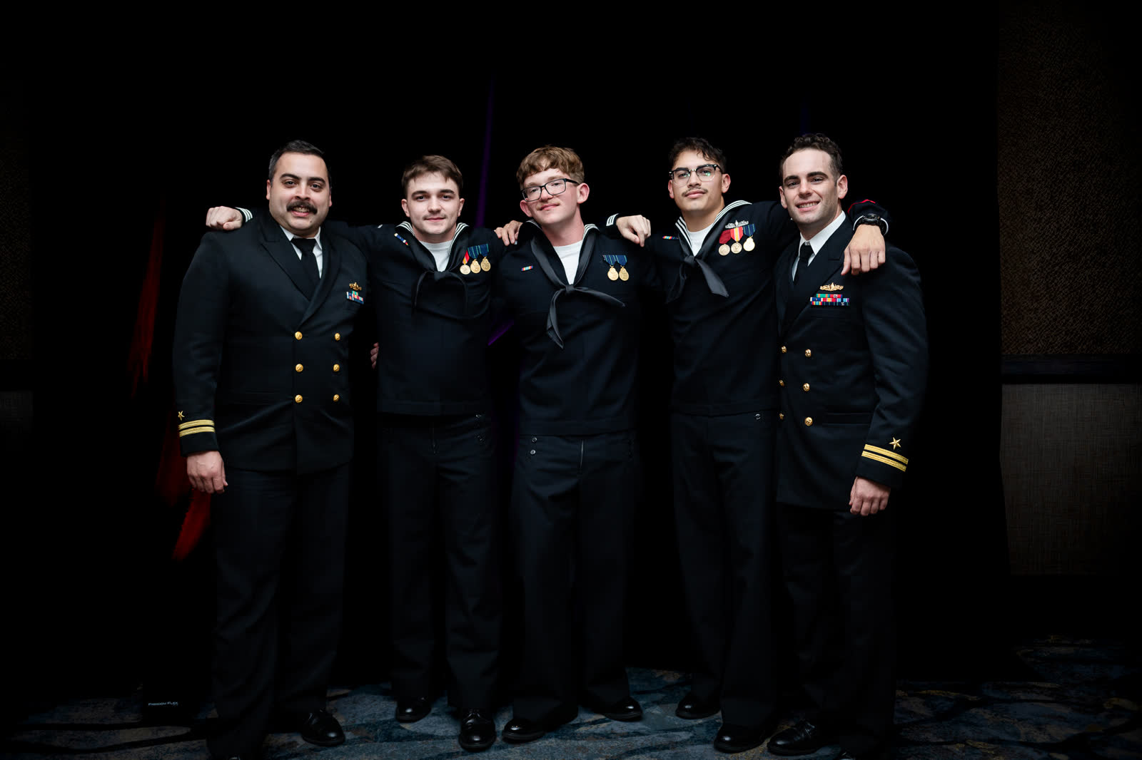 Five sailors and officers in dress uniform stand arm in arm for a portrait at the Navy Ball.