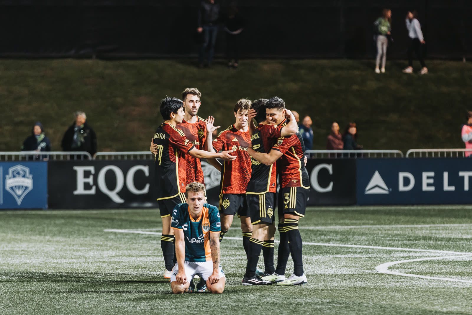 Sounders players celebrate together while an opposing player kneels on the turf.