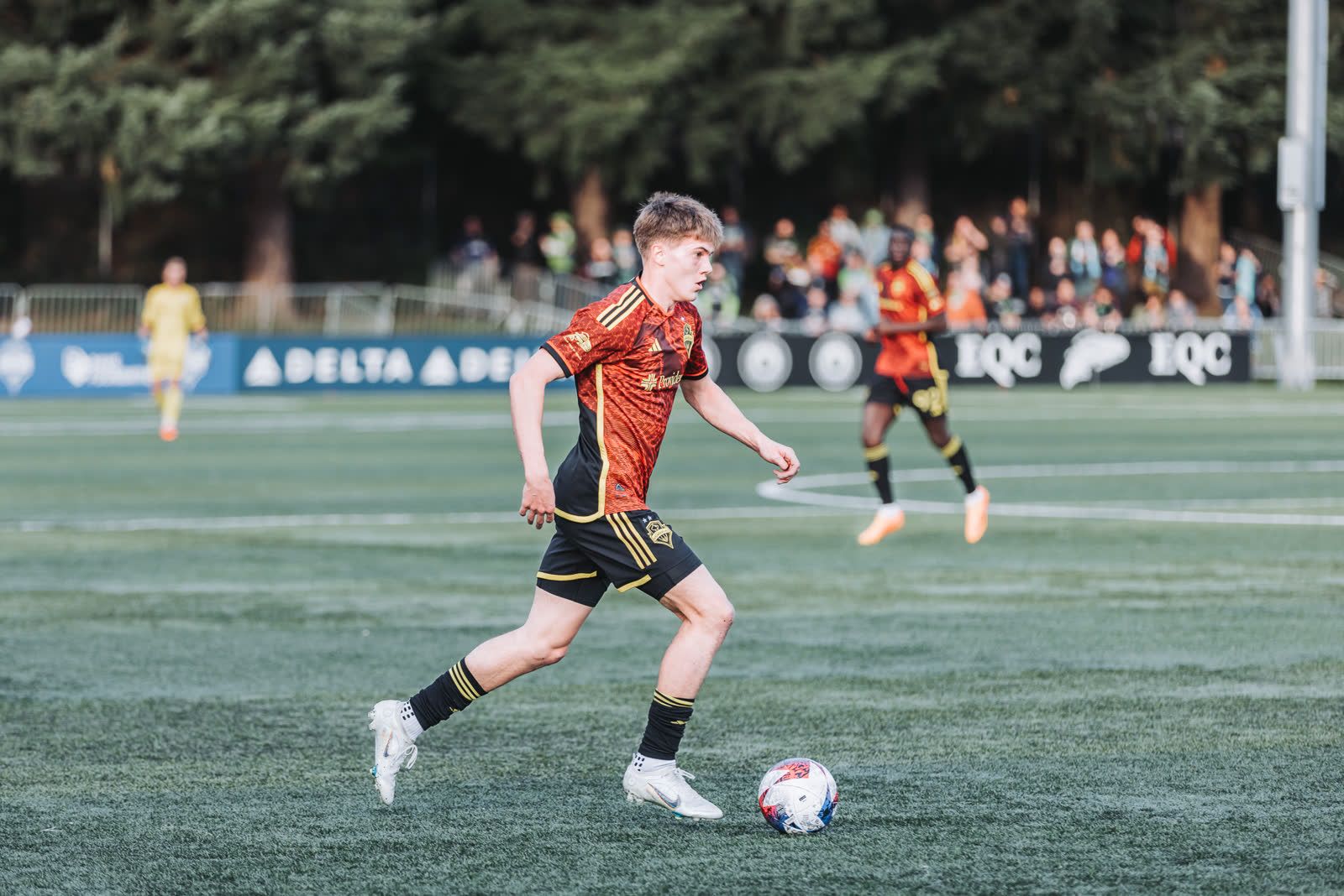 A Sounders player drives forward with the ball as the crowd watches from behind the boards.