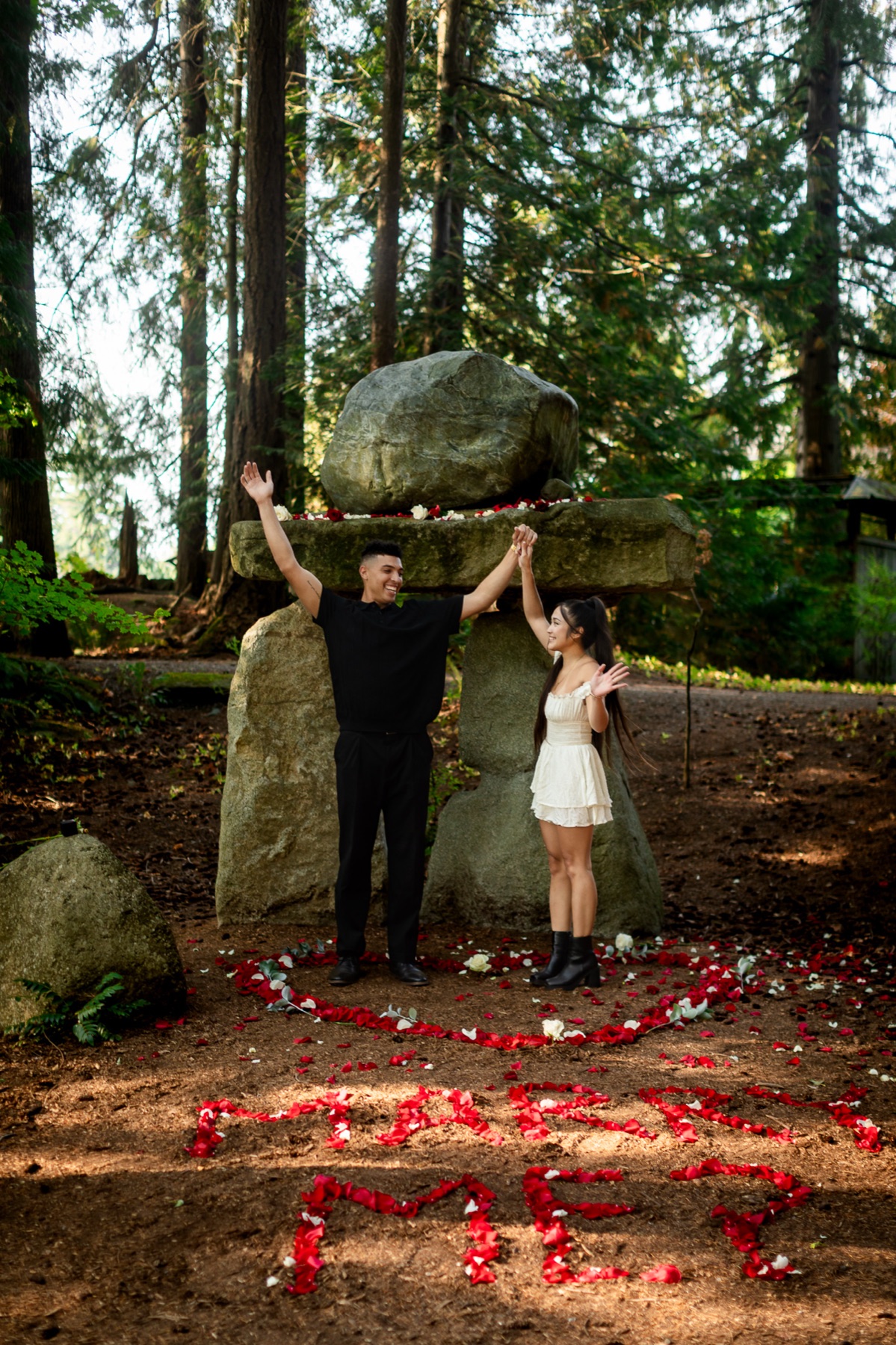 Trevor throws his arms up in celebration beside Jocelyn beneath the cedars, petals still on the ground.