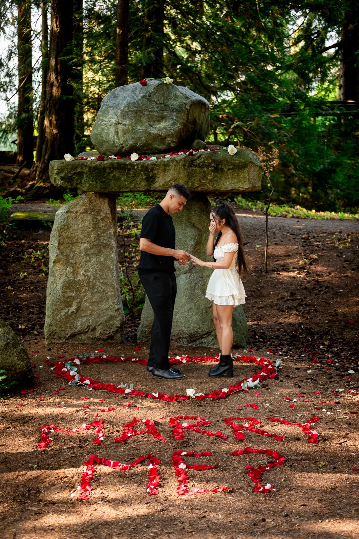Trevor slides the ring onto Jocelyn's hand under the stone arch, MARRY ME spelled out in petals below them.